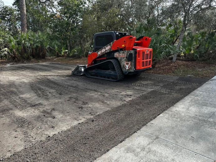 Red skid-steer loader on dirt beside a concrete path, with trees in the background.