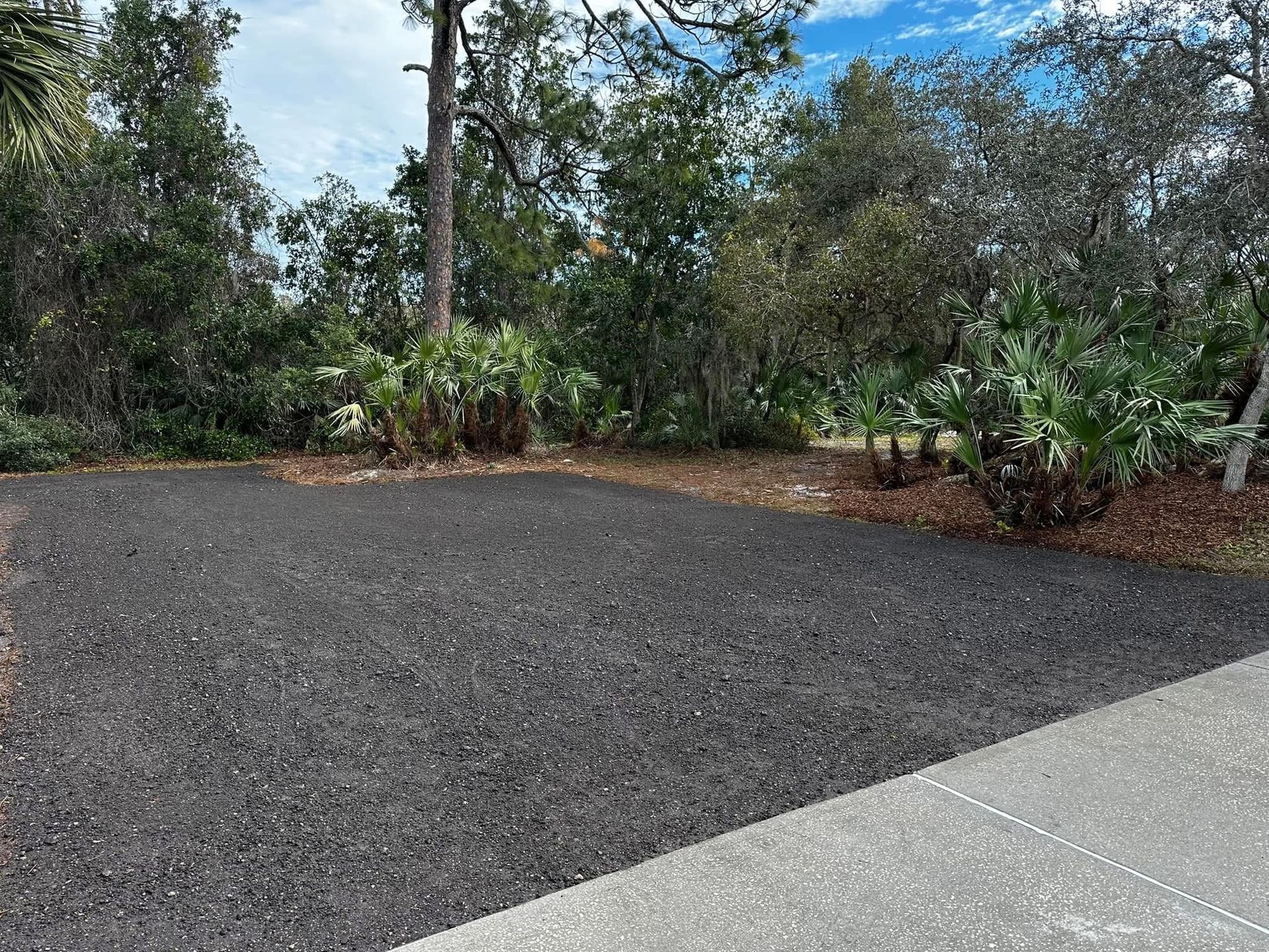 Gravel driveway beside a concrete path, bordered by trees and shrubs under a blue sky