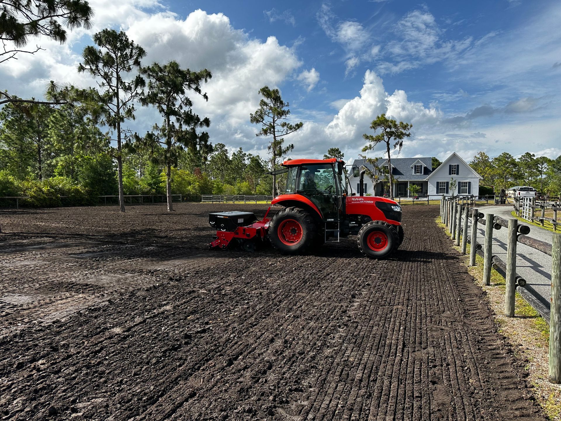Red tractor grading a muddy field beside a white fence under a partly cloudy sky
