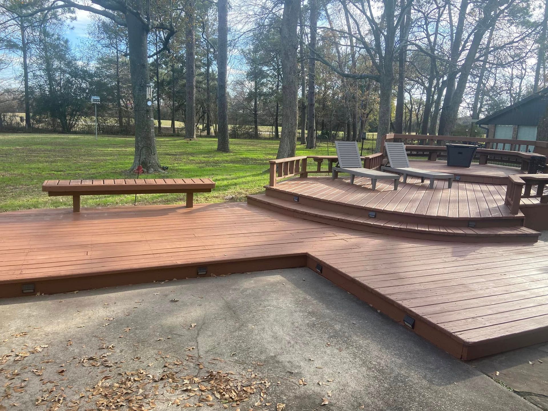 Wooden deck with curved steps and benches in a park, surrounded by trees and grass.
