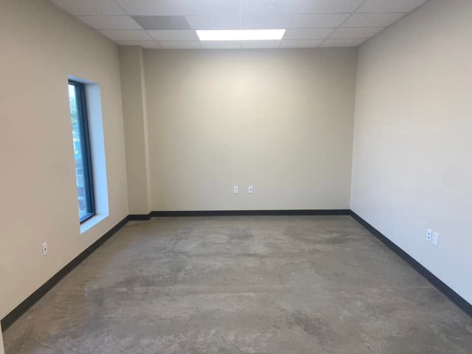 Empty beige office room with gray carpet, white ceiling tiles, and a narrow window on the left.