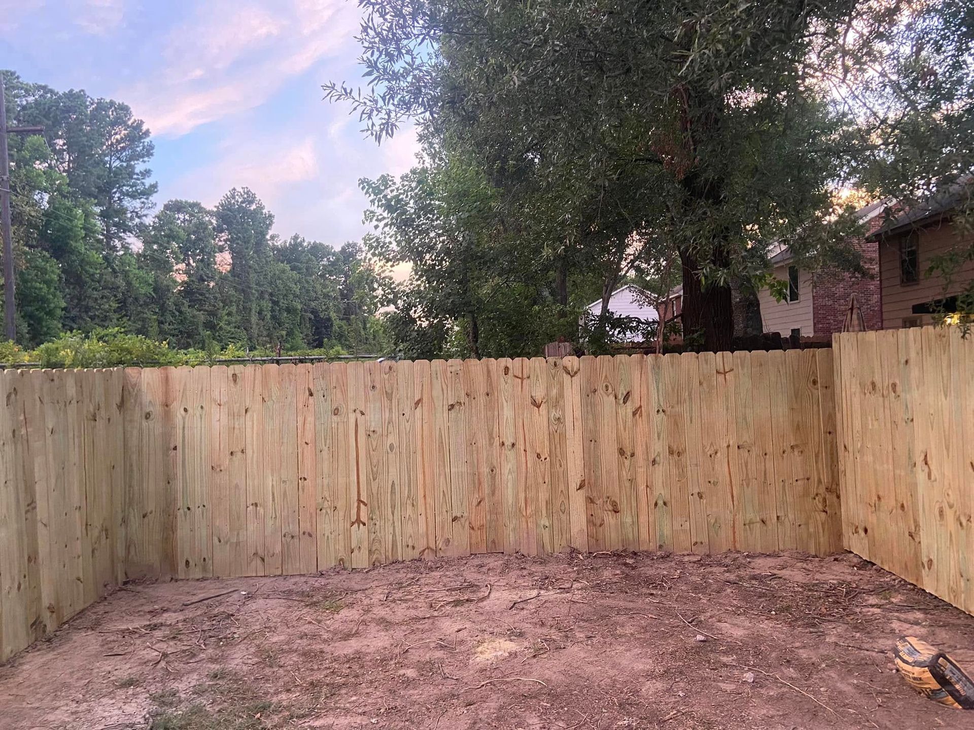 Backyard with a wooden privacy fence, dirt ground, and trees under a pink evening sky