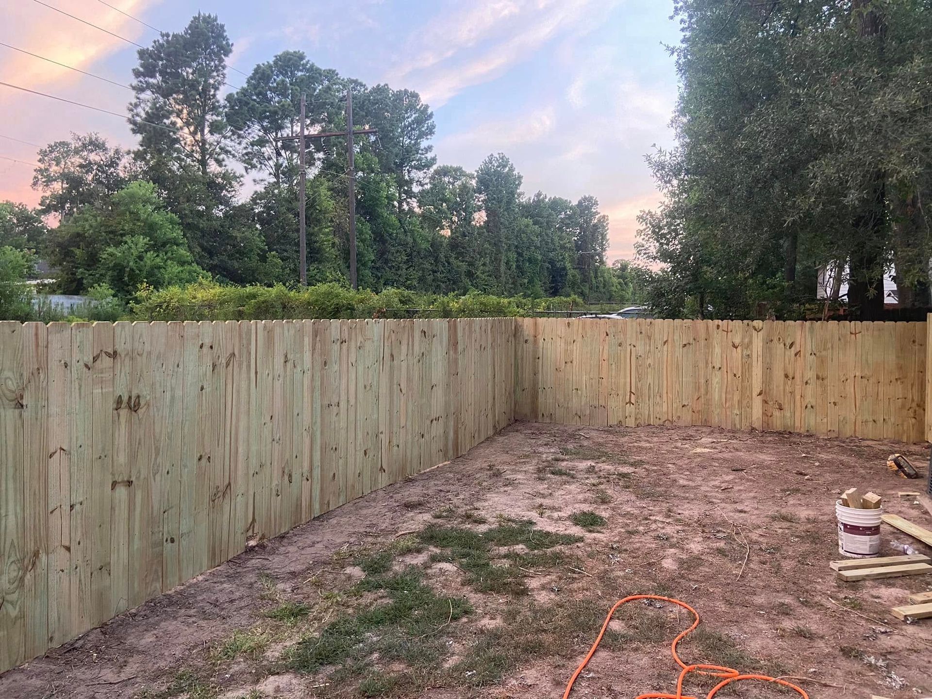 Freshly fenced backyard with bare dirt, patchy grass, and trees at sunset