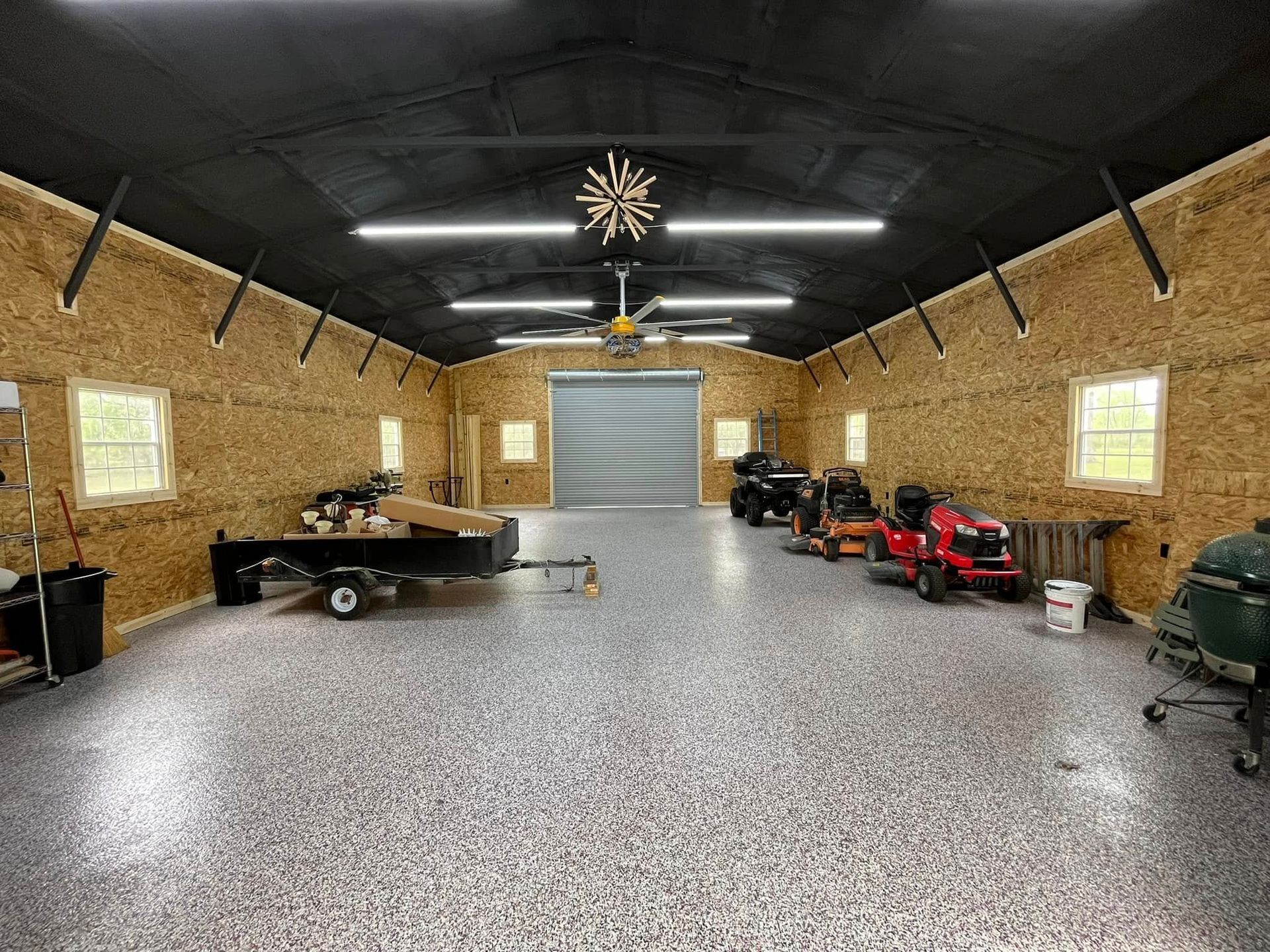 Wide garage interior with speckled floor, exposed wood walls, and several parked vehicles/tools.