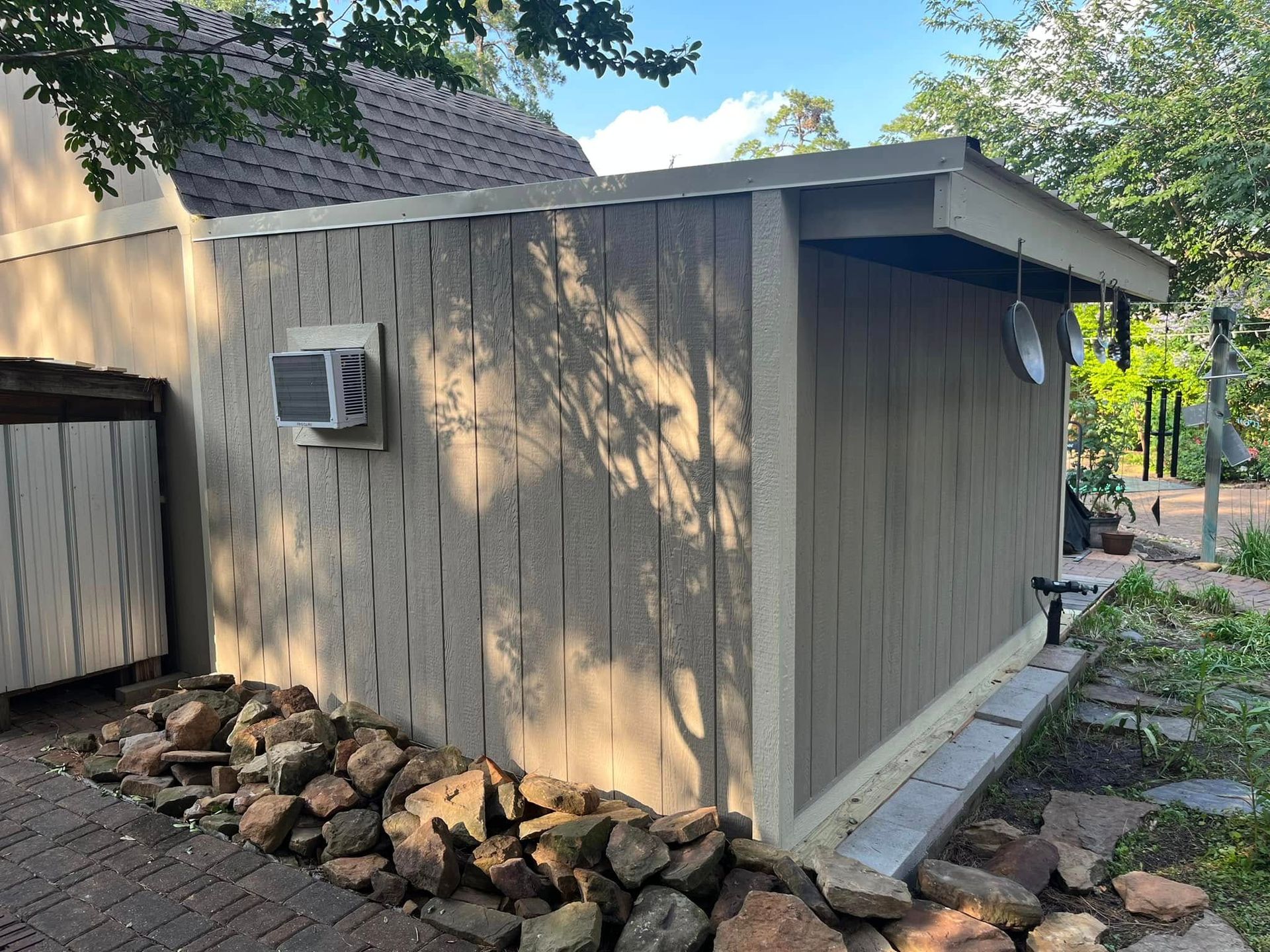 Tan wooden shed with a window AC unit, rock border, and stone path in a backyard area