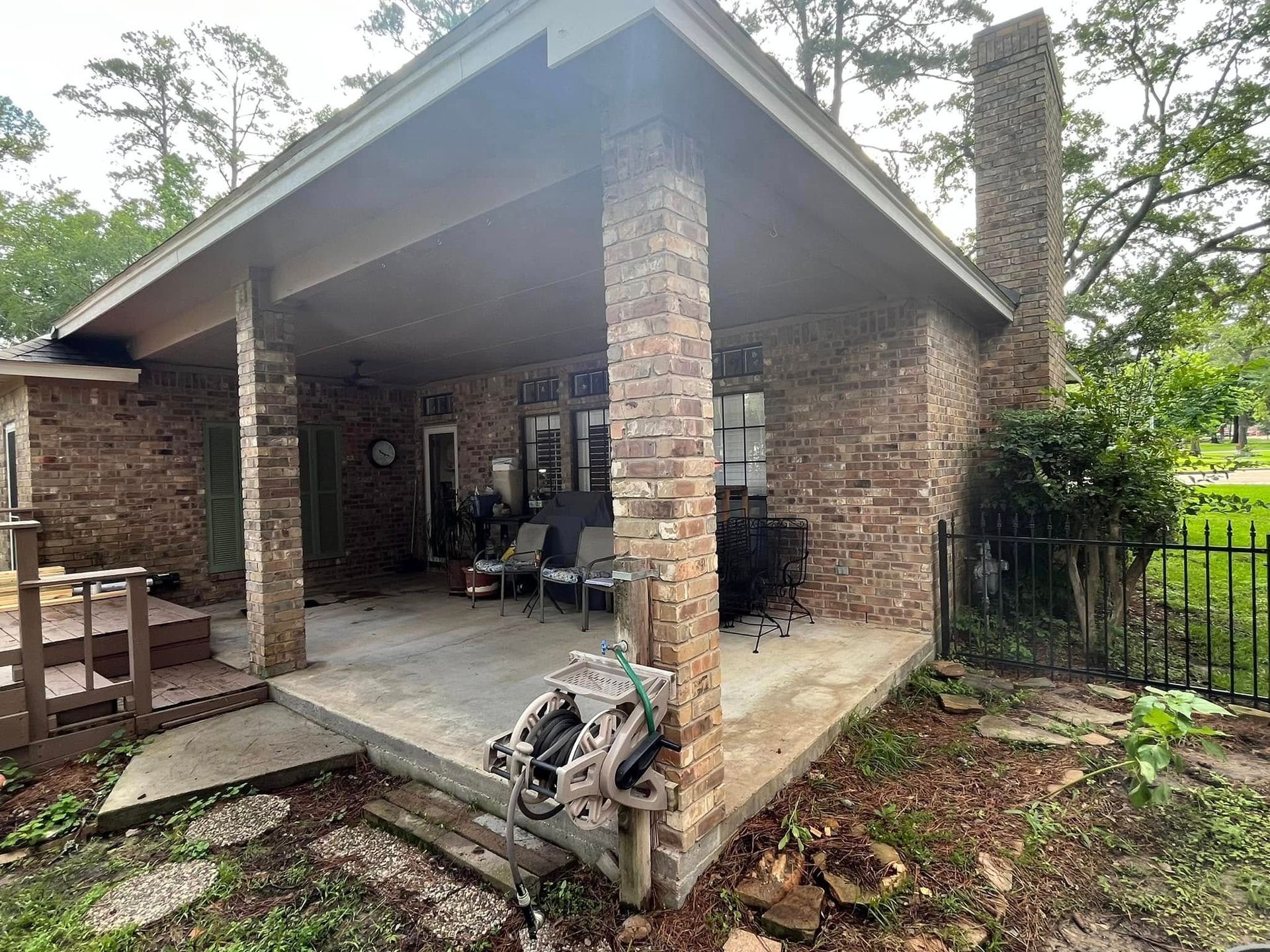 Brick house porch with chairs, potted plants, and tools in a yard under a sloped roof.
