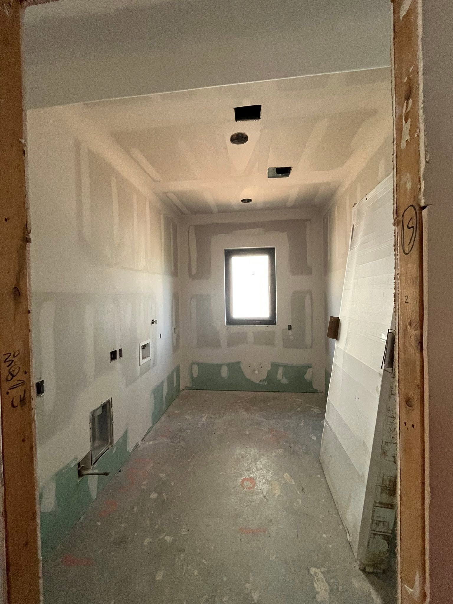 Narrow unfinished hallway under construction with drywall, exposed openings, and a small window at the far end