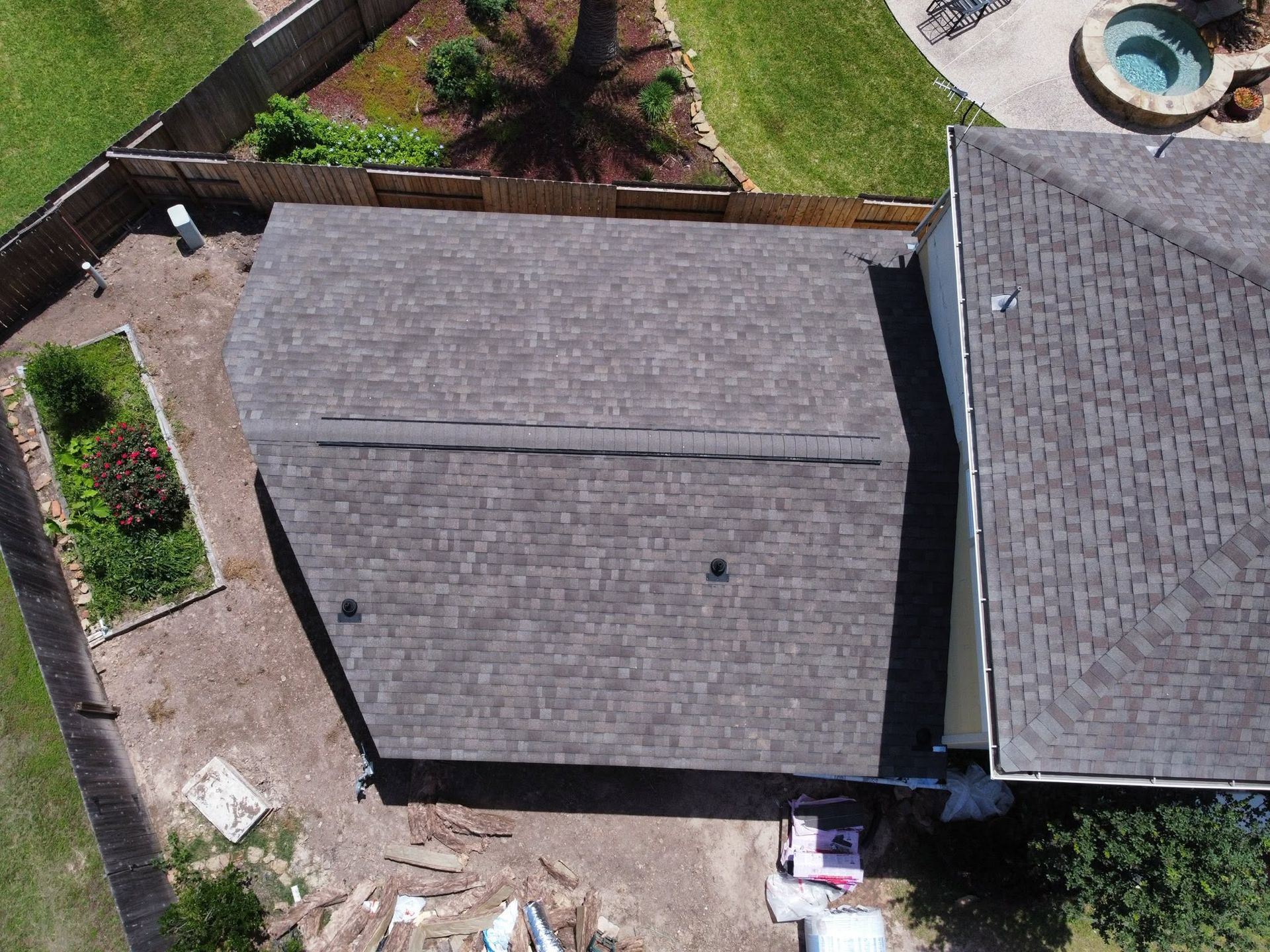 Aerial view of two gray-roofed houses with a backyard, fence, and small patio area.