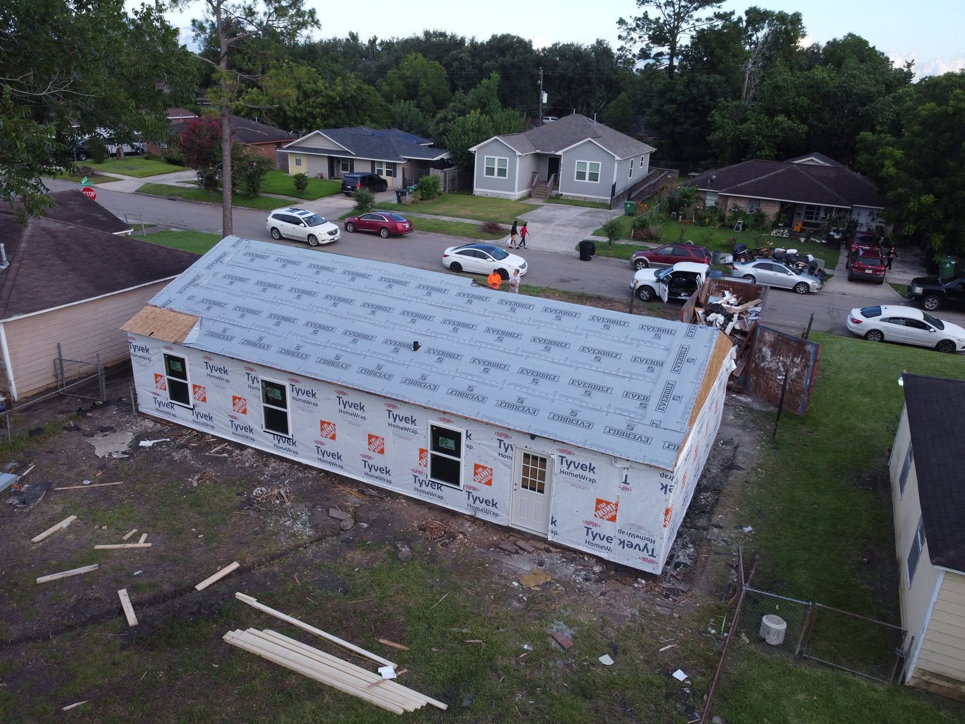 Aerial view of a house under construction with white roof wrap and several parked cars on a residential street.