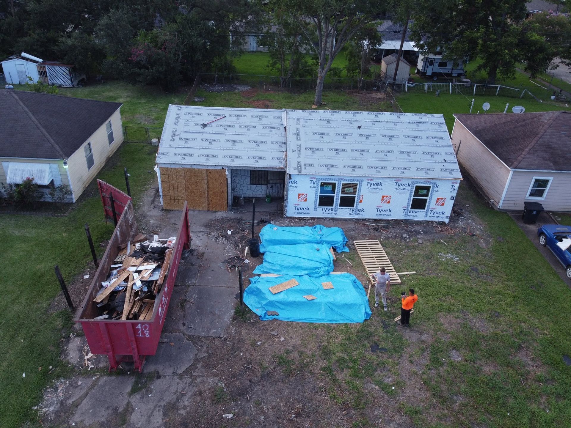 Aerial view of a house under renovation with blue tarps, a dumpster, and a worker in an orange vest.