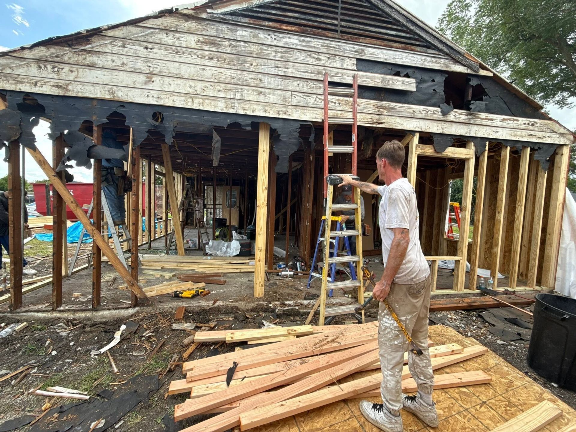 Person standing beside a partially demolished house with exposed wooden framing and debris.