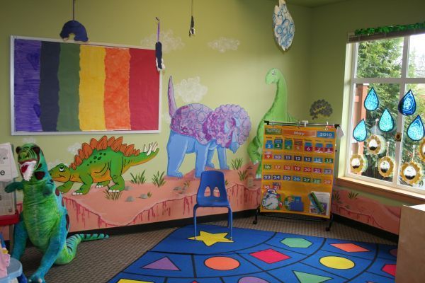 Colorful preschool classroom with rainbow flag, dragon mural, easel, rug, and windows with hanging decorations