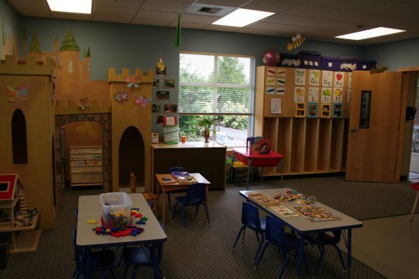 Bright preschool classroom with tables, toy shelves, cubbies, and a playhouse by a window