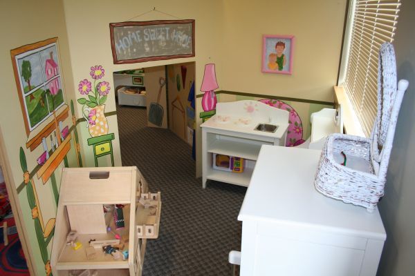 Child’s playroom with dollhouse, toy kitchen, white dresser, and wall art in a beige room.