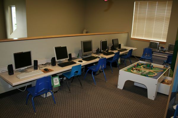 Empty computer lab with desktop stations and blue chairs beside a small play area.