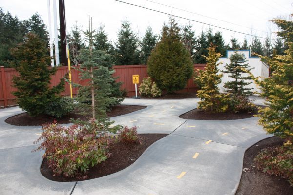 Curved paved path through landscaped shrubs and trees in a parking lot, with red fencing and a sign in the background