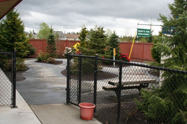 Chain-link fence entrance to a wet patio with picnic tables, red planter, and green signs under cloudy skies