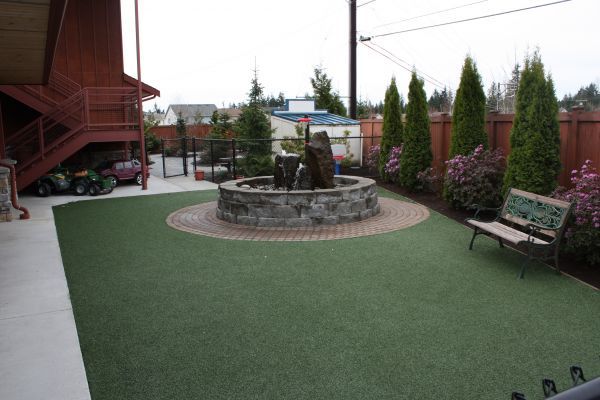 Courtyard with artificial turf, stone fountain, benches, and a wooden stairway beside a red building