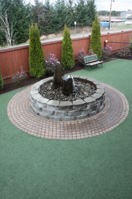 Circular stone planter with a small tree on a green artificial turf patio, beside a fence and bench
