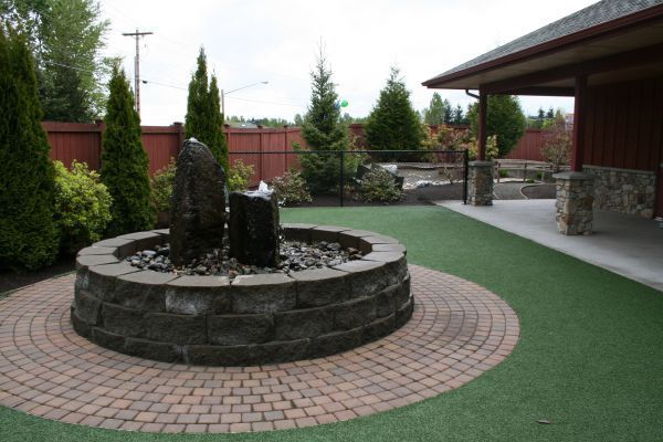 Circular stone fountain in a landscaped courtyard beside a covered building walkway.