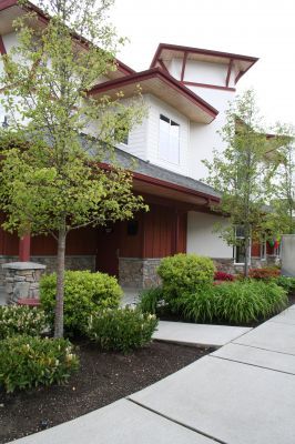 Modern house exterior with white walls, red roof accents, trees, shrubs, and a concrete walkway