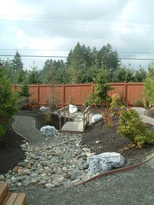 Landscaped backyard with a stone stream, small bridge, and red wooden fence under a cloudy sky