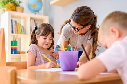 Teacher helping children draw at a classroom table with colored pencils and a purple cup