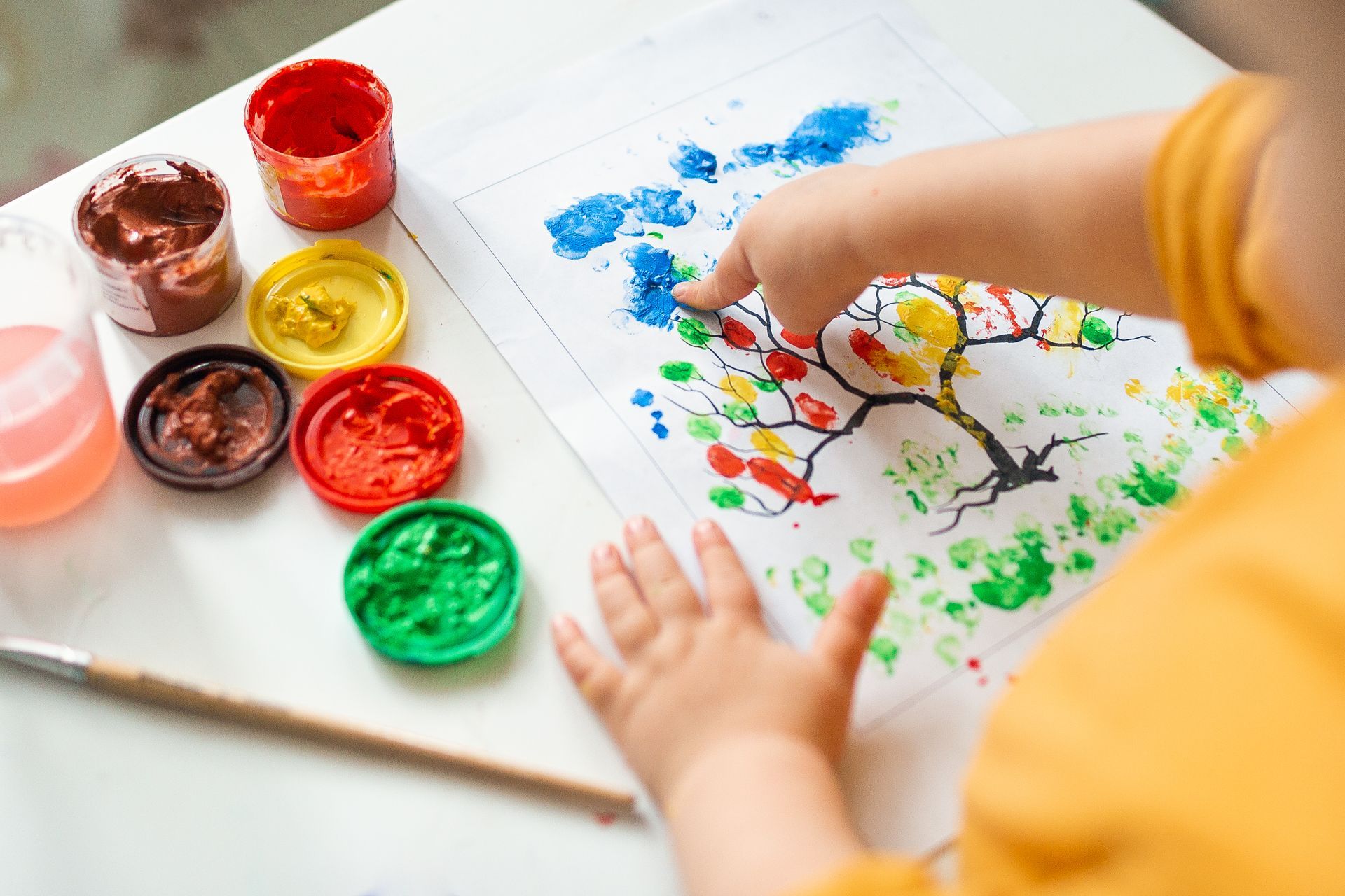 Child painting a tree with colorful finger paints on paper beside open paint pots