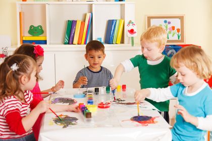 Children painting together at a table in a bright classroom, with colorful supplies and bookshelves behind them