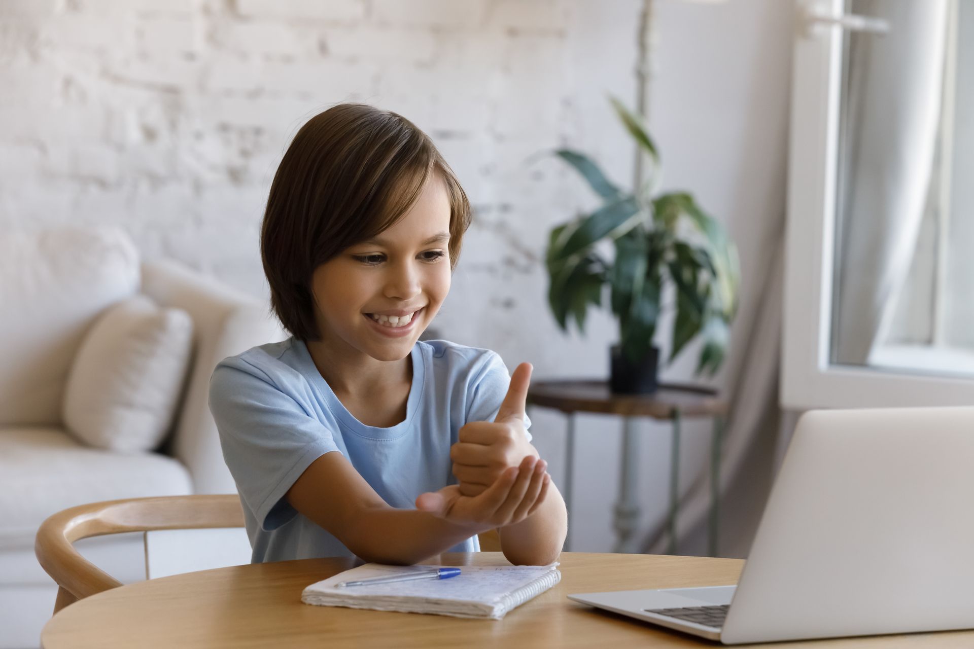 Smiling child giving a thumbs-up at a desk with a laptop and notebook in a bright room