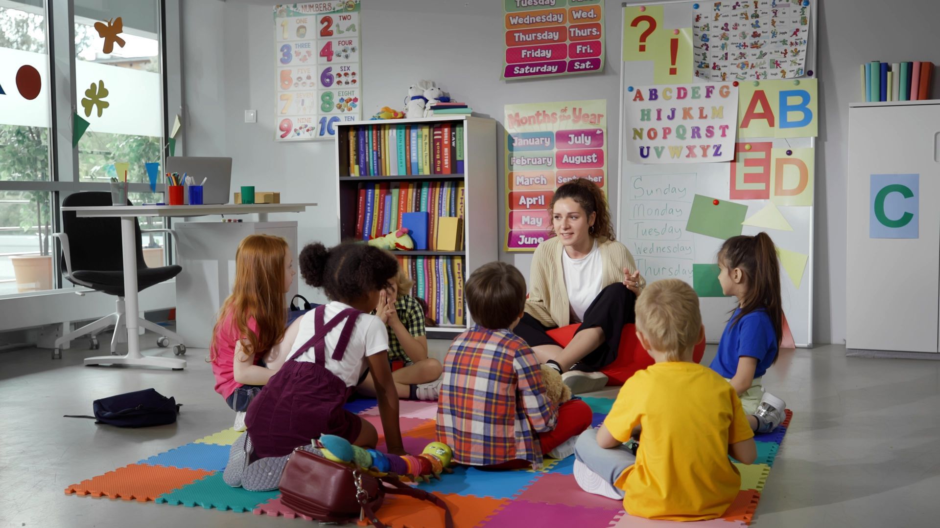 Teacher reading to children seated on colorful mats in a classroom with alphabet posters and bookshelves