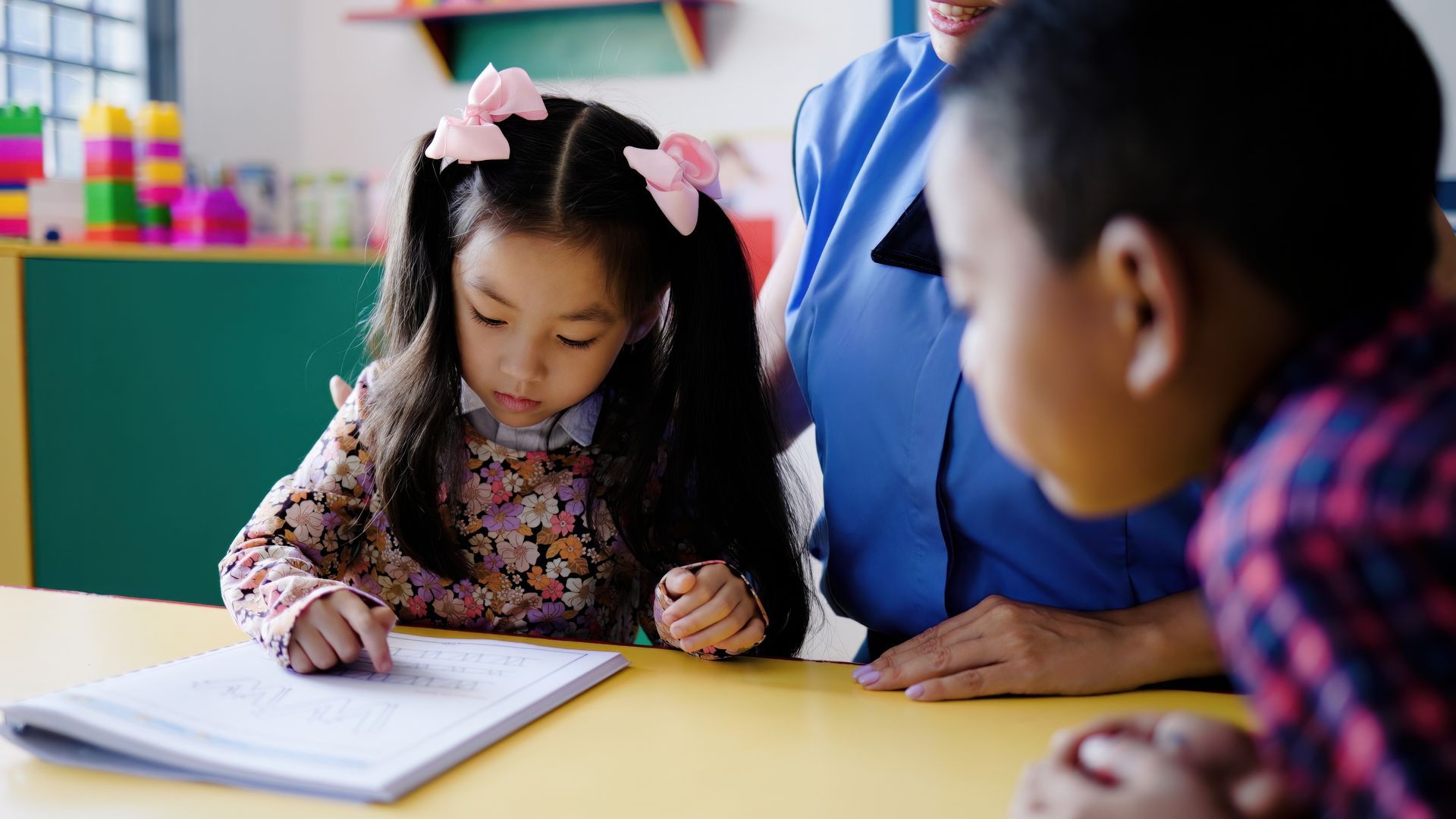 Child writing in a notebook with an adult nearby at a classroom table
