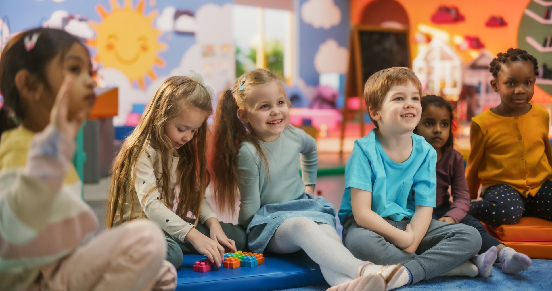 Children sitting on a blue mat and playing with toys in a colorful classroom, with an adult nearby