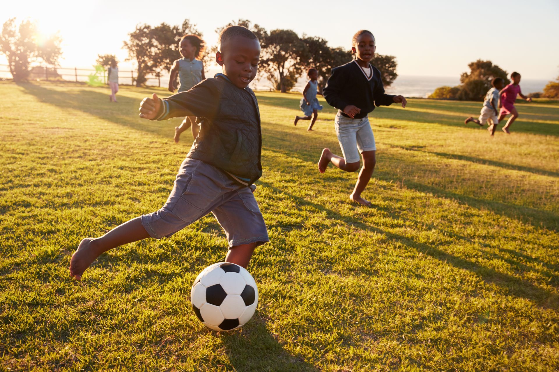 Kids playing soccer on a sunlit grassy field at sunset, with one kicking a black-and-white ball.