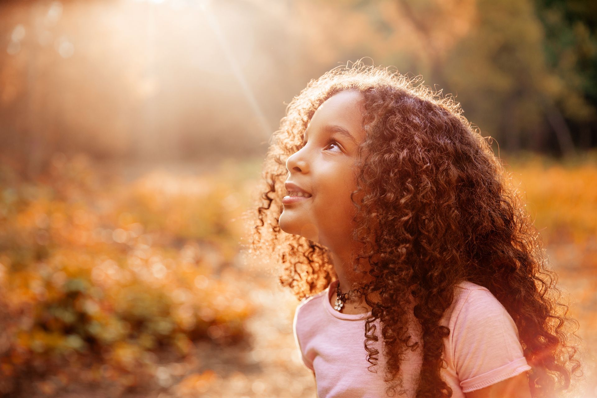 Woman with curly hair gazing upward in warm sunlight amid autumn leaves