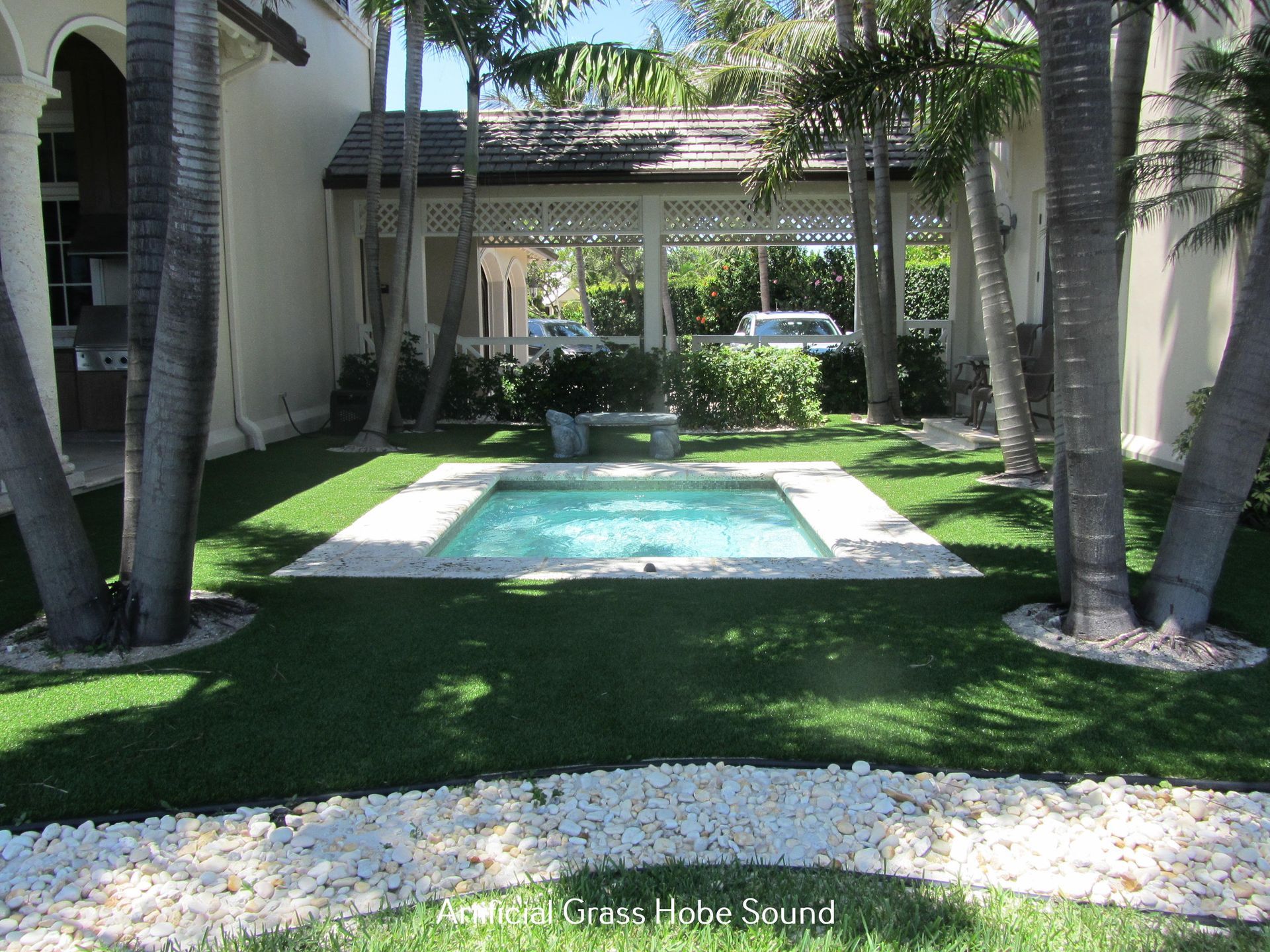 A small backyard with a pool, bordered by white stones and green grass, surrounded by palm trees.