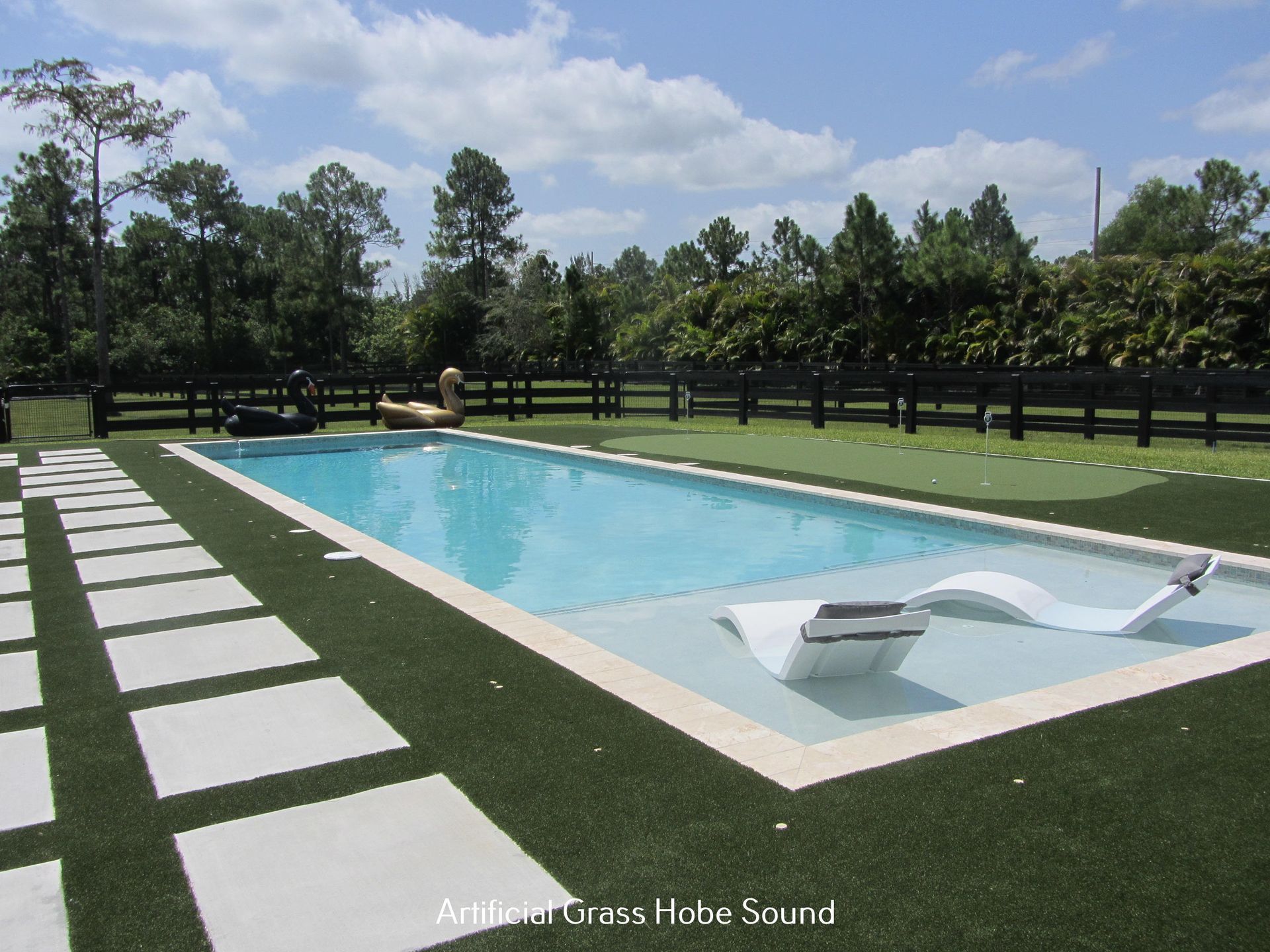 Rectangular pool with white lounge chair, surrounded by artificial turf and square stone pavers. Trees and a fence in the background.