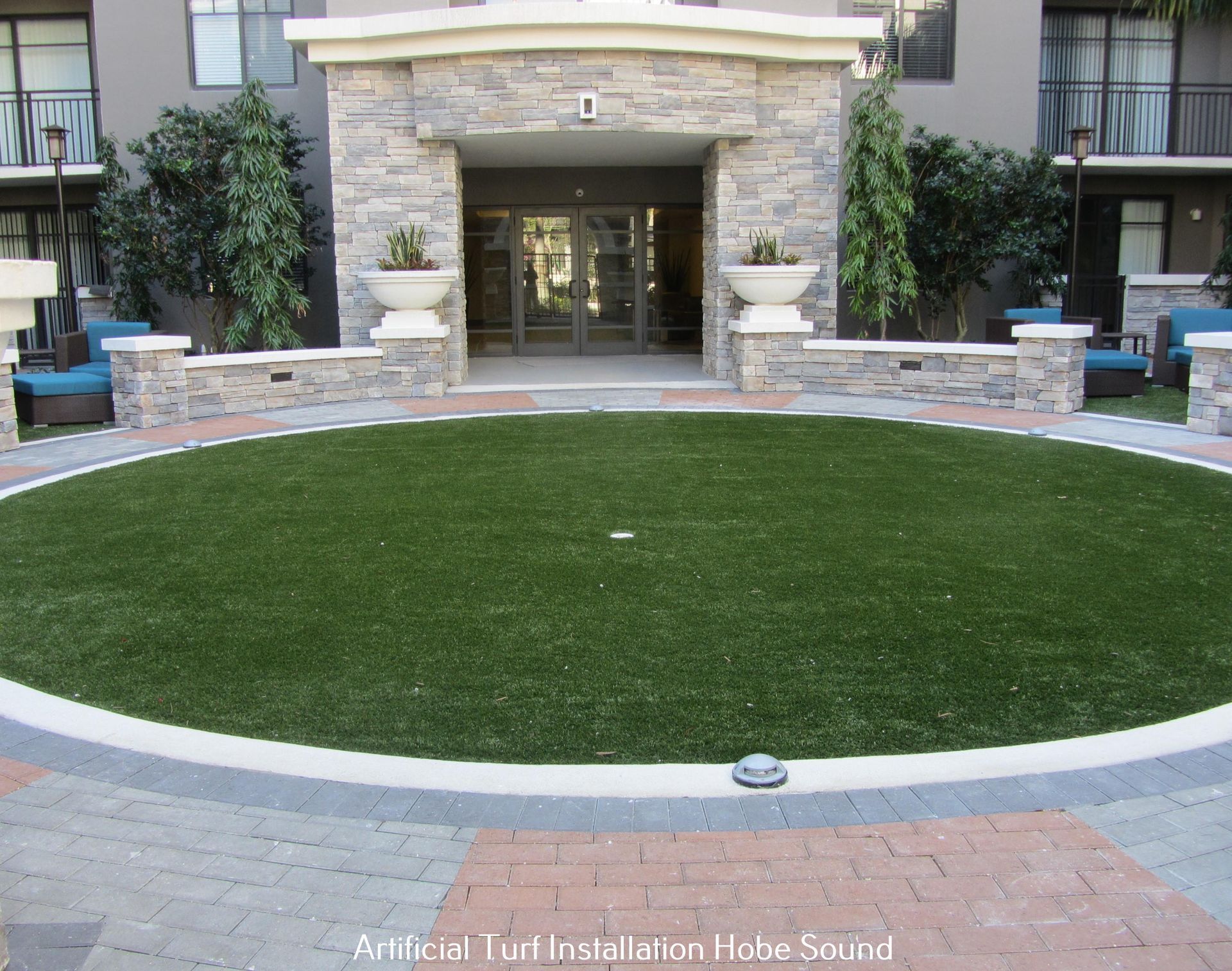 Circular artificial turf installation in front of an apartment building entrance, surrounded by brick and stone.