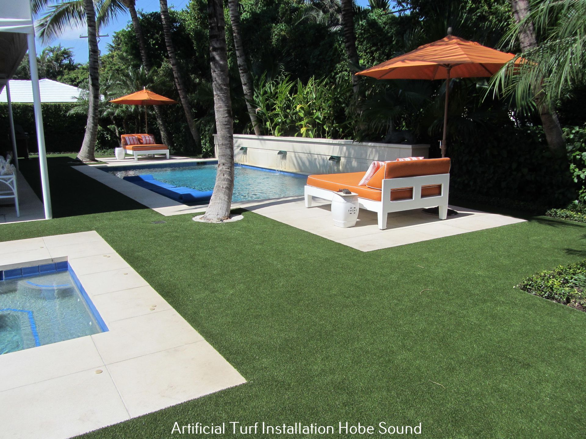 Poolside scene with artificial turf, lounge chairs, orange umbrellas, and a waterfall feature.