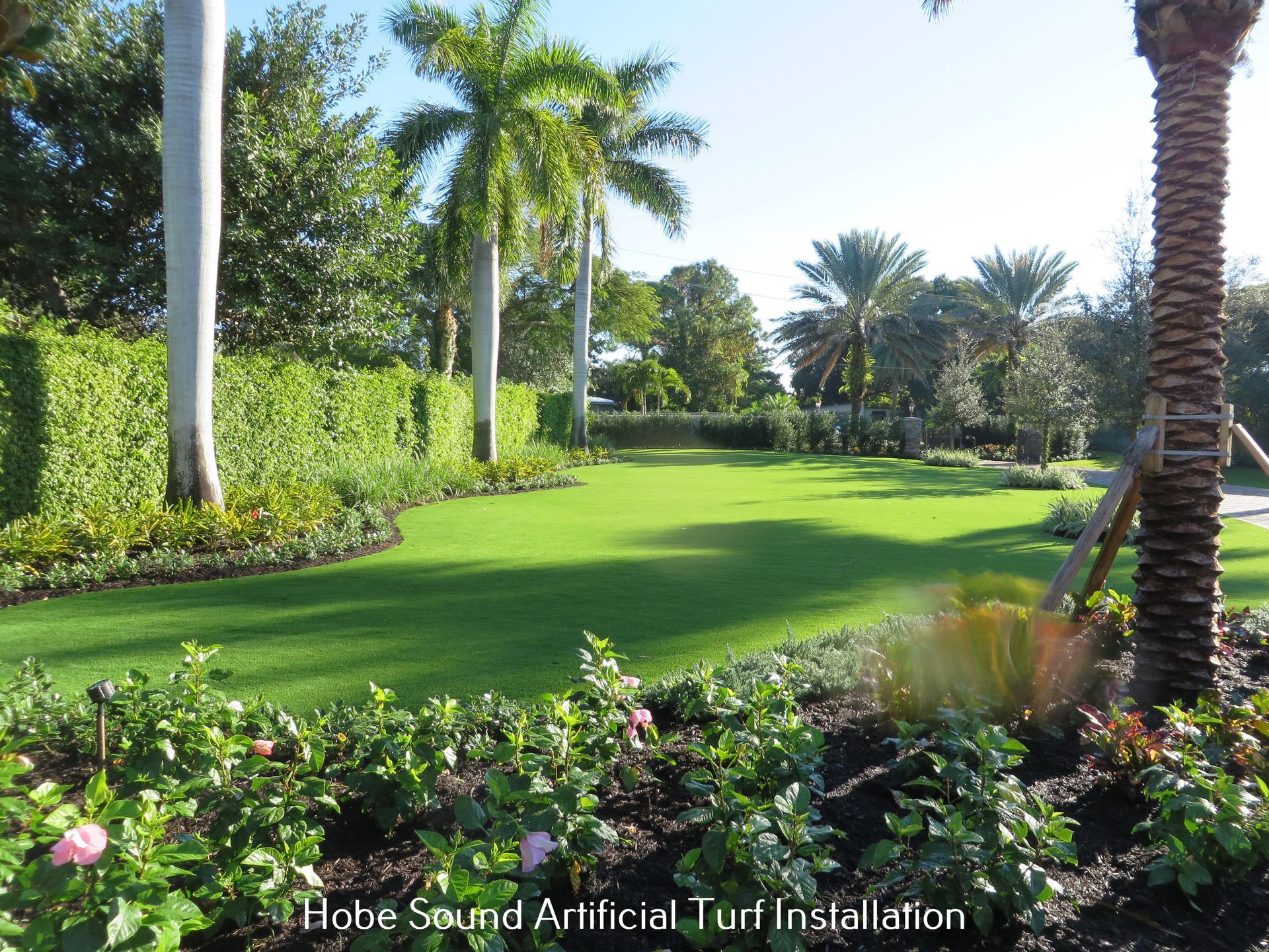 Lush green artificial turf lawn surrounded by trees and flowerbeds, sunny outdoor setting.