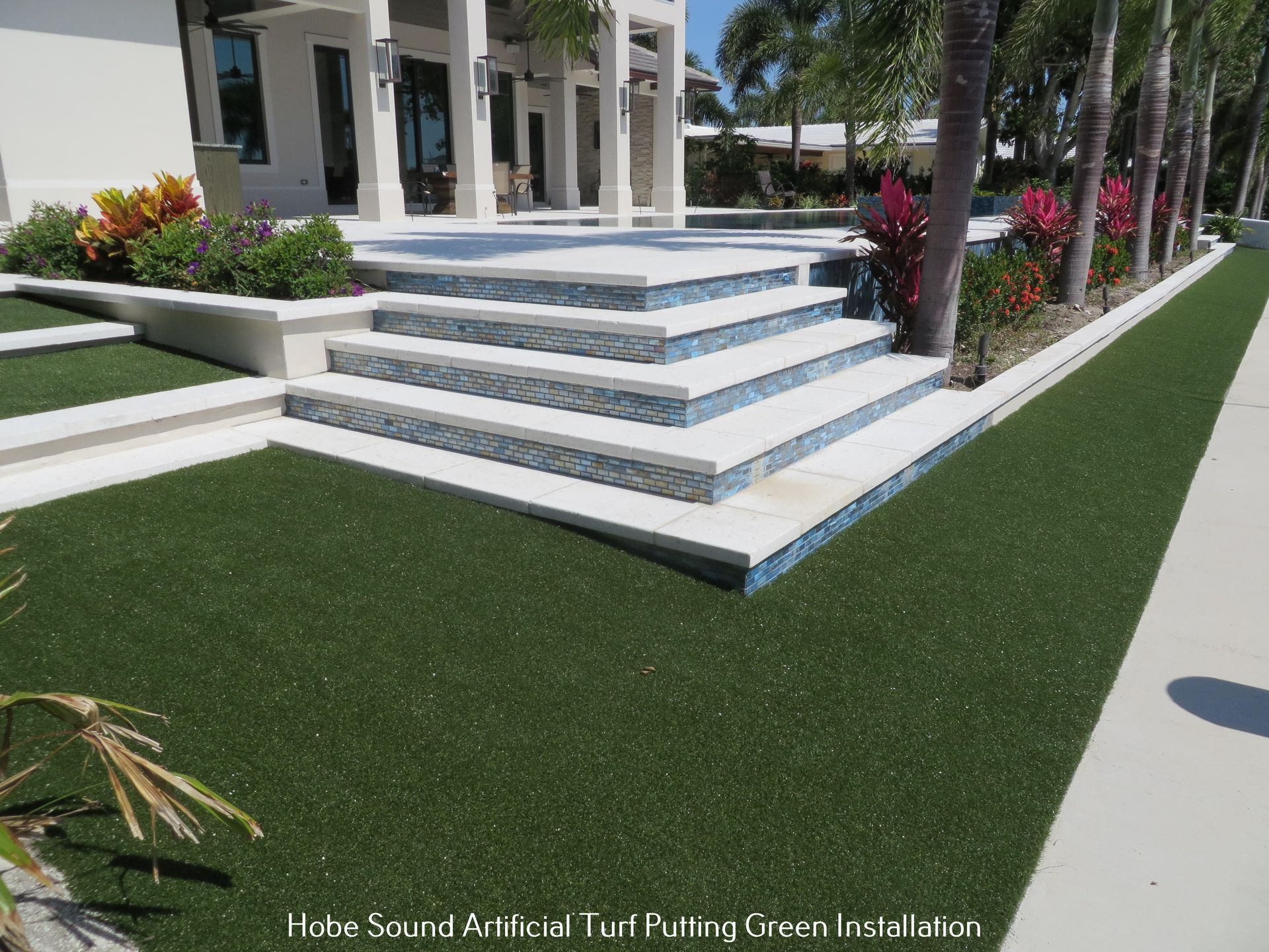 Artificial turf putting green installation next to a house with steps and a pool, palm trees in the background.