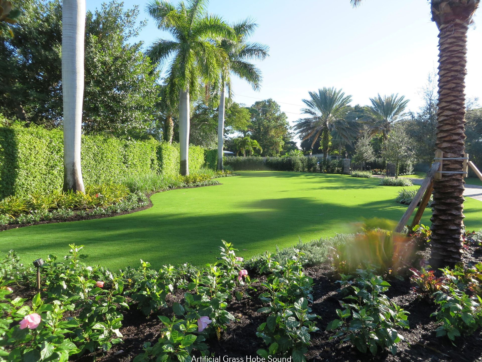Lush green lawn surrounded by manicured flower beds, palm trees, and hedges on a sunny day.