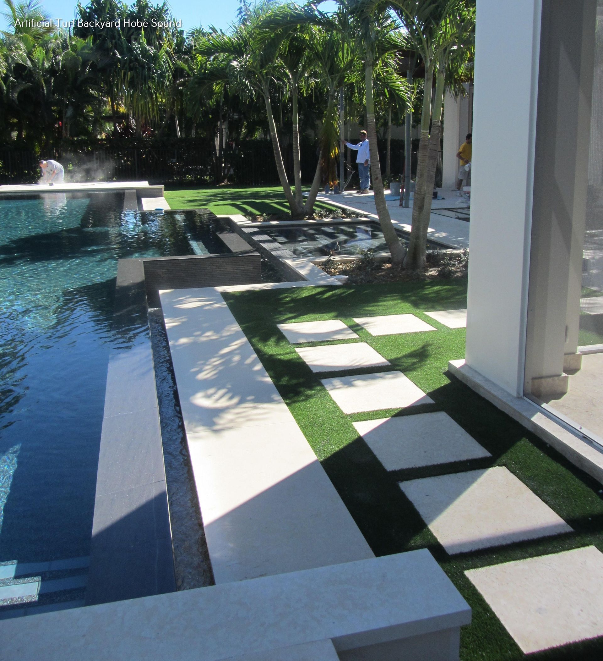 Poolside patio with square stepping stones, synthetic grass, and palm trees.