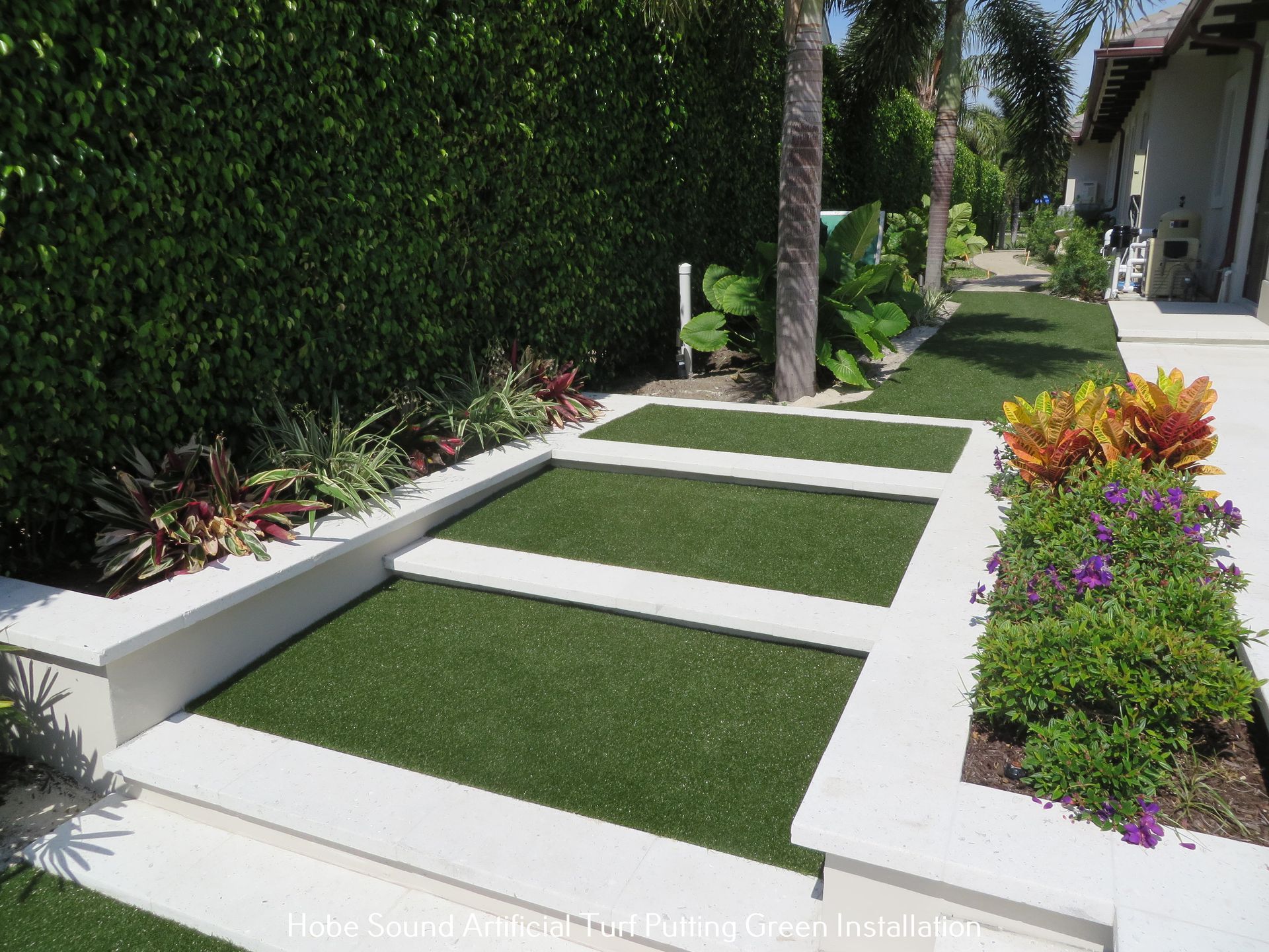 Artificial turf pathway with white borders, bordered by green hedge and flower beds.