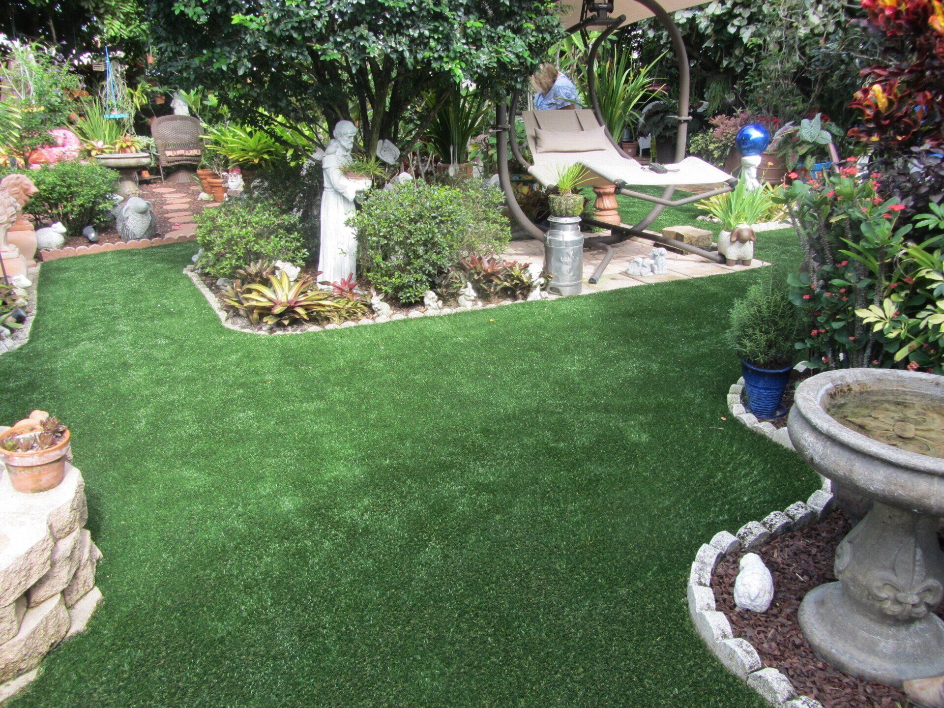 A lush, green garden with a stone statue, hammock, and birdbath.