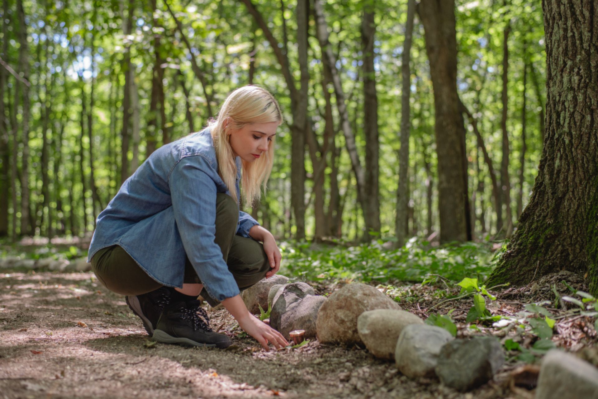 woman kneeling on a memorial forest path reaching out to touch a small metal marker 