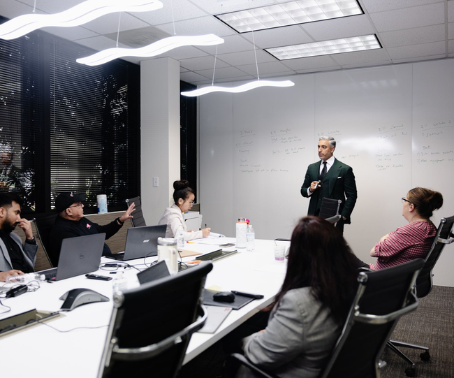 A man in a suit gives a presentation to a team in a modern office, using a whiteboard.