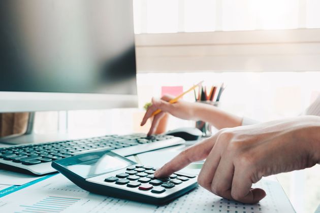 Hands using a calculator on a desk beside a keyboard and monitor in a bright office