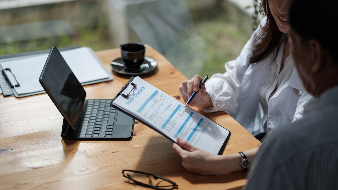 Woman pointing at document on clipboard, sitting with person near laptop and coffee cup.