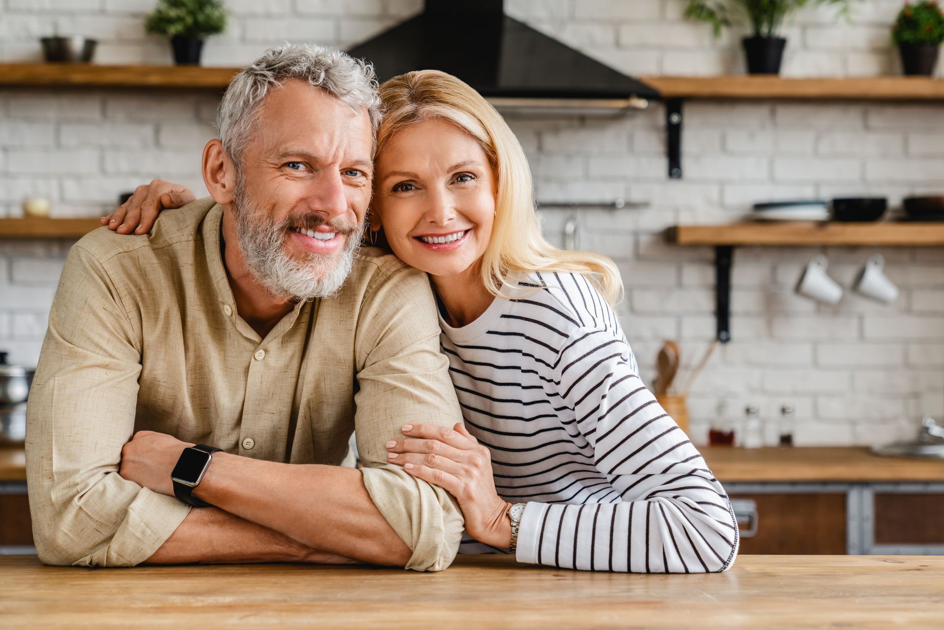 Smiling older couple in a kitchen, man with gray beard, woman with blonde hair, arm around each other.